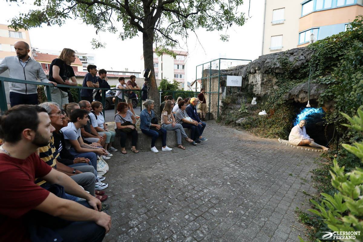 Une foule est assise en demi-cercle autor de Clélia qui est à l'entrée d'une petite grotte, elle même entourée de chanterelles blanches et de sculptures blanches de renards